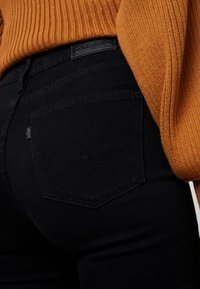 Jeans en denim noir avec un design de poche arrière incurvé, associés à un pull en maille côtelée orange texturé. Les jeans arborent une étiquette logo sur la poche.