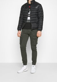 Czarna kurtka puchowa z zamkiem błyskawicznym z przodu, czarny t-shirt z grafiką pod spodem, oliwkowe spodnie bojówki z bocznymi kieszeniami oraz białe trampki.