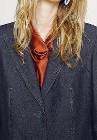 Denim blejzr v tmavě modré barvě s oranžovým prošíváním, s jedním knoflíkem na zapínání, strukturovanými rameny, vrstvený přes hedvábnou blůzu v rezavé barvě.