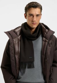 Sort uldblandingshal med frynser i enderne, båret over en grå sweater og en quiltet brun jakke, der viser et lagdelt vinterlook.