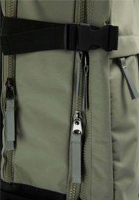 Gros plan sur l'avant d'un sac à dos vert avec plusieurs fermetures éclair et une sangle noire réglable avec boucle en plastique.