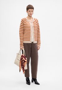 Cardigan a righe arancione e crema, top beige, pantaloni marroni, stivali neri con tacco, che tiene una borsa crema con una sciarpa decorata legata sopra.