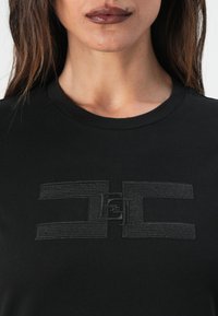 Svart T-skjorte med en teksturert, tonet logo over brystet. Stoffet er glatt og har et klassisk rundhalsdesign.