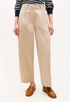 Person trägt beige weit geschnittene Hosen, gestreiften marineblau-weißen Pullover und braune Lederloafers und steht vor einem schlichten Hintergrund.