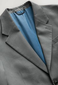Grau Blazer mit Reverskragen, ausgestattet mit einem hellblauen Futter und einem dekorativen Kettendetail. Ein-Knopf-Verschluss und glatte Textur.