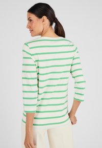 Vrouw met donker haar in een paardenstaart, gekleed in een wit en groene horizontaal gestreepte blouse met lange mouwen en lichtgekleurde broek, met haar rug naar de camera gekeerd.