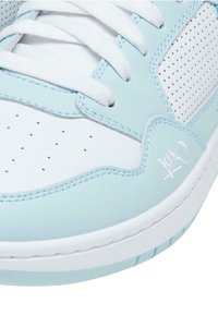 Sneaker azzurro chiaro e bianco con una parte superiore in sintetico liscio, pannelli laterali perforati, lacci bianchi piatti e dettagli del logo in rilievo.
