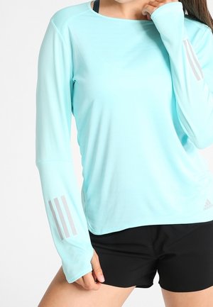 Kobieta w jasnoniebieskiej sportowej bluzce z długim rękawem z odblaskowymi paskami na rękawach oraz w czarnych szortach, pozująca z ręką przy twarzy.