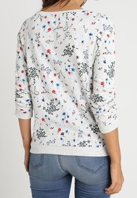 Suéter de manga larga gris con estampado floral y pequeños patrones en rojo, azul y verde. Textura suave, escote redondo y mangas ajustadas.