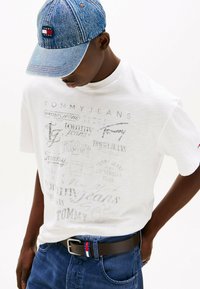 Camiseta blanca con estampados gráficos en gris deslavado, gorra de denim azul, cinturón marrón con acentos en rojo, blanco y azul, y jeans azules.
