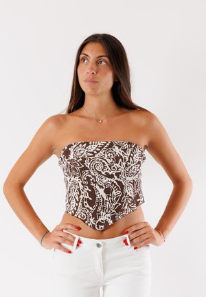 Una giovane donna si erge con sicurezza indossando un top senza spalline marrone e bianco a fantasia, abbinato a jeans bianchi, con le mani sui fianchi.