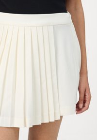 Pleated skirt in off-white fabric, featuring a wide waistband and stitched hem. The fabric has a smooth texture, enhancing its structured design.