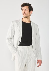 Hvid stribet habitjakke med en enkelt knap, kombineret med en sort t-shirt. Stof ser glat ud; moderne snit med reverskrave.