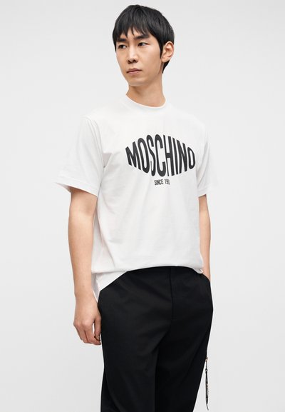 Άσπρο βαμβακερό μπλουζάκι με μαύρο λογότυπο "MOSCHINO" και το κείμενο "SINCE 1983", συνδυασμένο με μαύρο φαρδύ παντελόνι, φορεμένο casual.