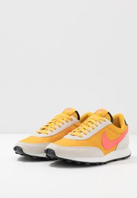 Nike-sneakers med ett gult och rosa färgschema, med överdel i mocka och tyg, svarta accenter och en texturerad svart gummisula.