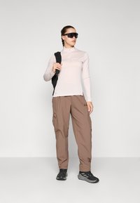Light pink long-sleeve top with a zip collar, paired with brown cargo pants featuring pockets, and black hiking shoes. Black backpack and sunglasses.