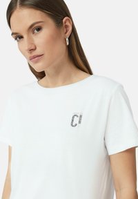 Weißes T-Shirt mit rundem Ausschnitt und kurzen Ärmeln, versehen mit einem taschengroßen Logo im schwarz-weißen Leopardenmuster auf der Brust.
