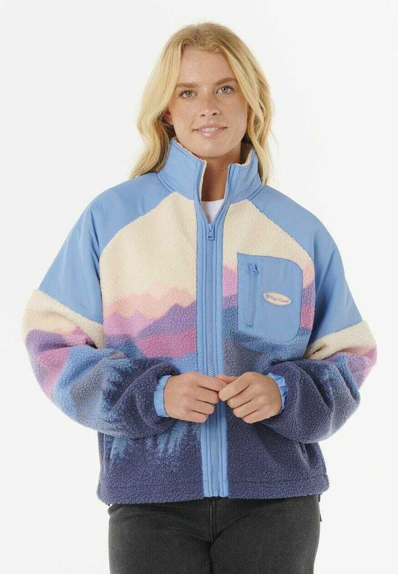 Kvinne med blonde hår iført en fargerik fleecejakke i blå, lilla, rosa og kremfargede toner, stående foran en hvit bakgrunn.