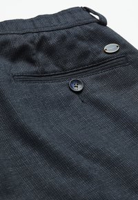 Tissu à carreaux gris foncé avec une poche arrière boutonnée et une petite étiquette de marque métallique ovale près de la ceinture.