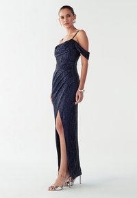 Večerna obleka temno modre barve z bleščicami, z izrezom čez ramena, stranskim razporkom in oprijetim krojem. Nošena s srebrnimi sandali z visokimi petami.