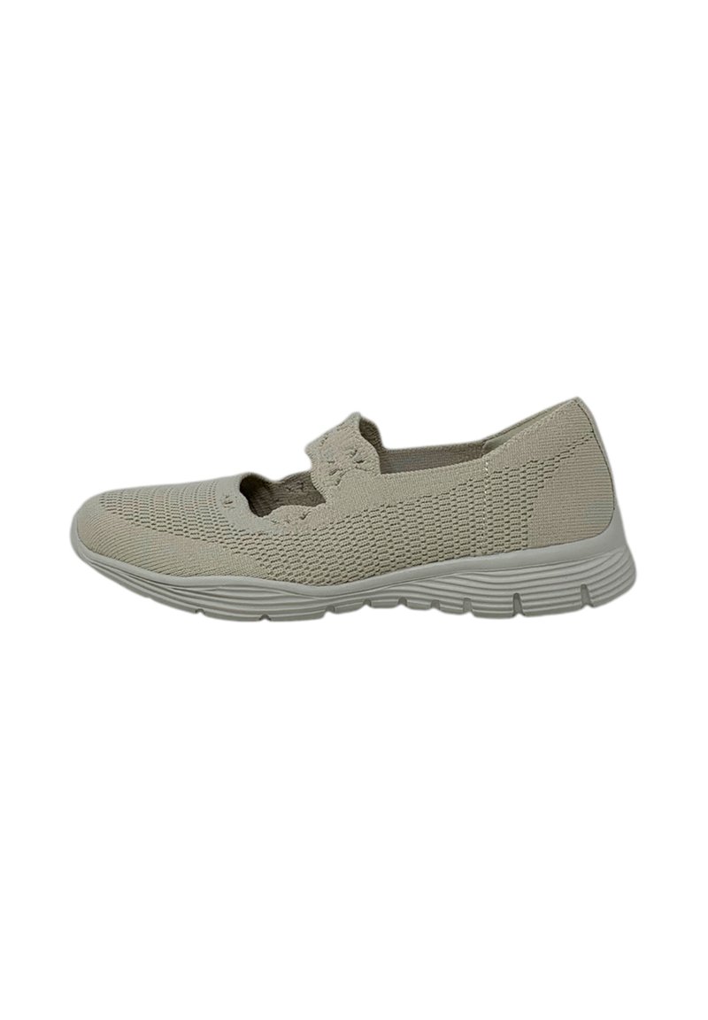 Zapato beige tipo slip-on con tejido de punto, lados abiertos y suela blanca flexible. Presenta una superficie texturizada y un sutil detalle de patrón.