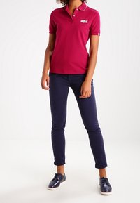 Fuchsia poloshirt met kraag en Lacoste-logo, gecombineerd met een navy fitted broek en glanzende blauwe schoenen. Eenvoudig en strak ontwerp.