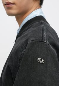 Fekete farmerdzseki bordázott gallérral, bal vállán ezüst logóval. Közeli kép a texturált anyagról és a varrás részleteiről.