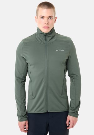 Mannelijk model dat een olijfgroene jas met rits en hoge kraag draagt, met het Vaude-logo op de borst, staand tegen een effen witte achtergrond.