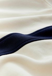 Krémová látka s vlnitým námorníckym modrým akcentom, ktorá má hladkú textúru a čisté stehy. Dizajn vytvára vizuálny kontrast a plynulosť.