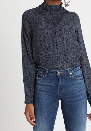 Langärmliges Oberteil in Marineblau mit strukturiertem Muster, hohem Rüschenausschnitt und lockerer Passform, kombiniert mit klassischen blauen Jeans.