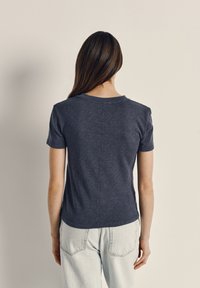 Woman with long straight hair stands facing a plain light wall, wearing a dark gray short-sleeve shirt and light blue jeans.