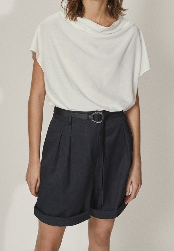 White draped short-sleeve top paired with dark navy high-waisted shorts featuring a belt with a circular buckle and rolled cuffs.