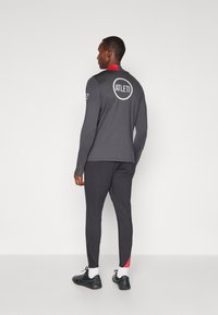 Sort langærmet træningsskjorte med en rød krave, som har et cirkulært "ATLETI" logo på bagsiden. Sammen med mørke bukser og sorte sneakers.