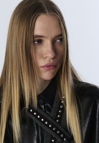 Giacca di pelle nera con borchie argentate e dettagli in tessuto grigio, con colletto classico e finitura testurizzata. Capelli lunghi e lisci.