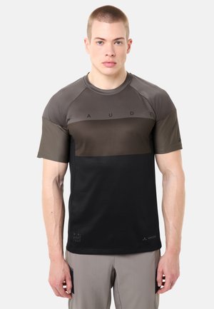 Jeune homme aux cheveux courts portant un t-shirt sportif à manches courtes gris et noir en blocs de couleur, et un pantalon gris clair, debout devant un fond blanc.