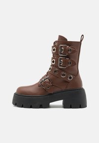 Brune faux skinn ankelstøvler med flere sølvspenne-detaljer, sirkulære øyer, stablet svart plattformsåle og sidehåndtak.