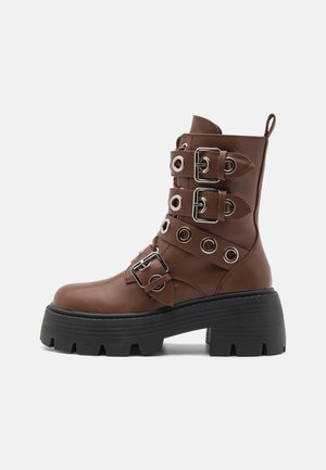 Brune faux skinn ankelstøvler med flere sølvspenne-detaljer, sirkulære øyer, stablet svart plattformsåle og sidehåndtak.