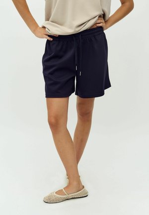 Navy blue athletic shorts with an elastic waistband and drawstring, featuring a loose fit and smooth texture. Worn with light-colored flat shoes.
