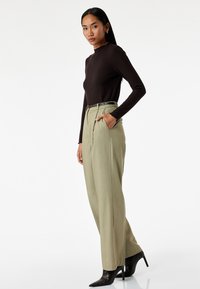 Donkerbruin geribbelde lange mouwen top gecombineerd met lichtgroene high-waisted broek, voorzien van plooien en een zwarte riem, aangevuld met puntige zwarte hakken.