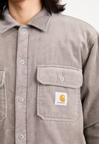 Grau korduroy Hemd mit einer Knopfleiste, zwei Brusttaschen und einem Carhartt-Logo-Patch. Strukturierter Stoff und hellgraue Knöpfe.