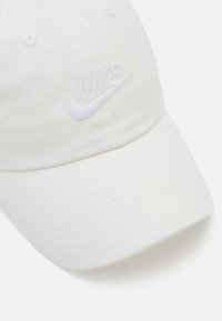 Balta medvilninė beisbolo kepuraitė su lenktu kraštu, ant priekinės dalies turi ryškų baltą siuvinėtą Nike logotipą. Tekstūruotas paviršius su siuvimo detalėmis.