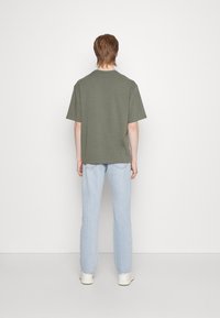 Olive green short-sleeve t-shirt with a straight fit, paired with light blue straight-leg jeans and white sneakers, viewed from behind.