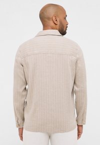 Camicia a maniche lunghe a righe beige realizzata in tessuto leggero. Presenta un colletto classico e polsini con bottone, con sottili righe bianche su una superficie testurizzata.