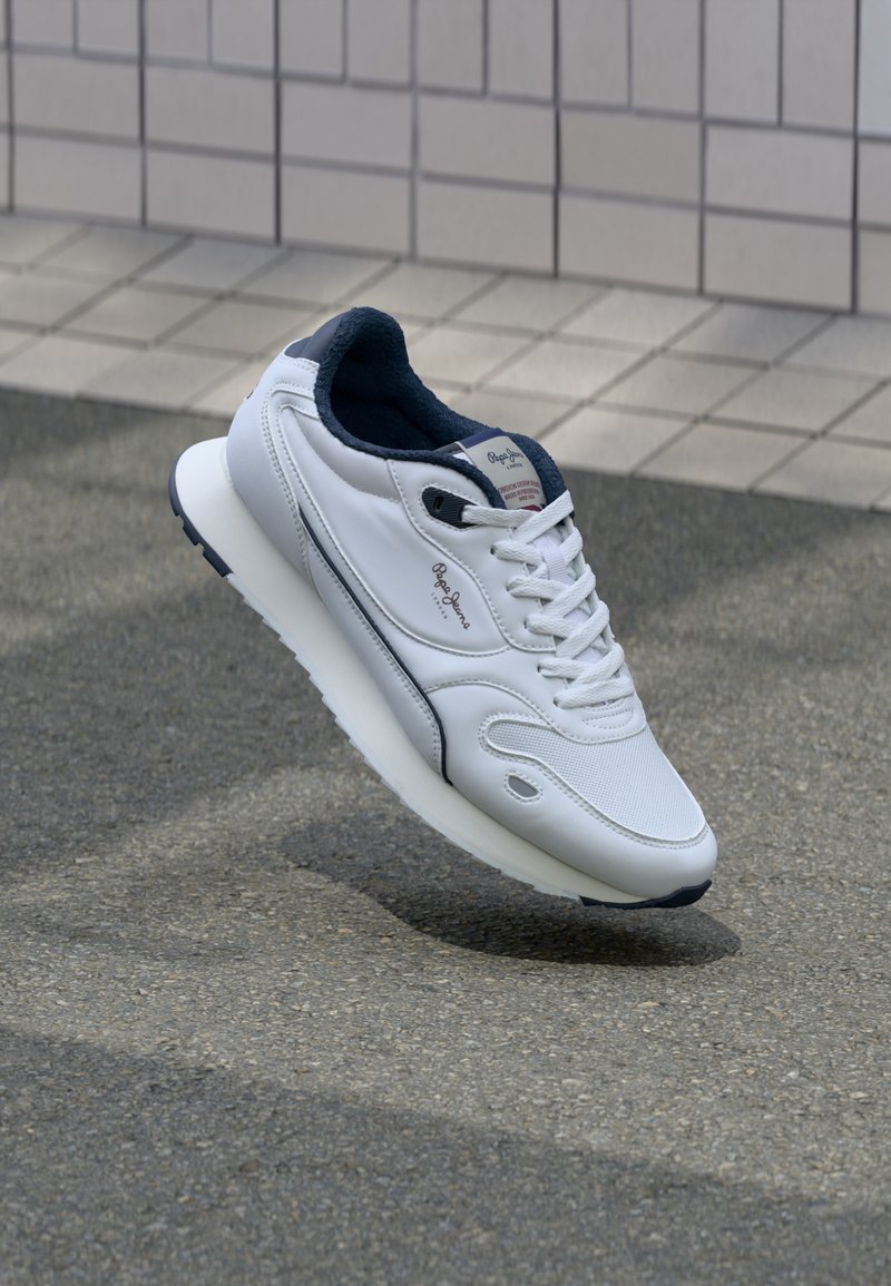 Baskets blanches avec une tige en cuir texturé, accents bleu marine, bout arrondi et semelle blanche rembourrée. Présente des lacets et un logo sur la languette.