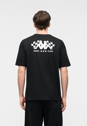 Άνδρας που φοράει μαύρο μπλουζάκι με άσπρο σχέδιο δύο καθιστών φιγούρων να κρατούν σημαίες καρό και κείμενο "FONDATO *** IN ITALIA" στην πλάτη.