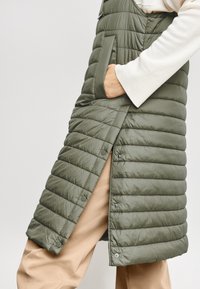 Hand i fickan på lång, olivgrön vadderad väst med tryckknappar över beige byxor och creamfärgad långärmad tröja.