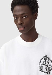 Valkoinen puuvillat-paita, jossa on musta luukuvio vasemmassa rinnassa, rento istuvuus ja pyöreä kaula-aukko.