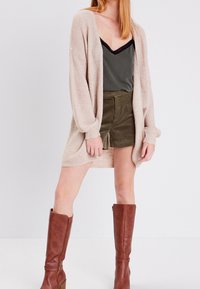 Stickad beige cardigan, lång ärm, öppen fram. Oljefärgad linne under. Oljefärgade shorts med snedställd fram. Bruna knähöga stövlar.
