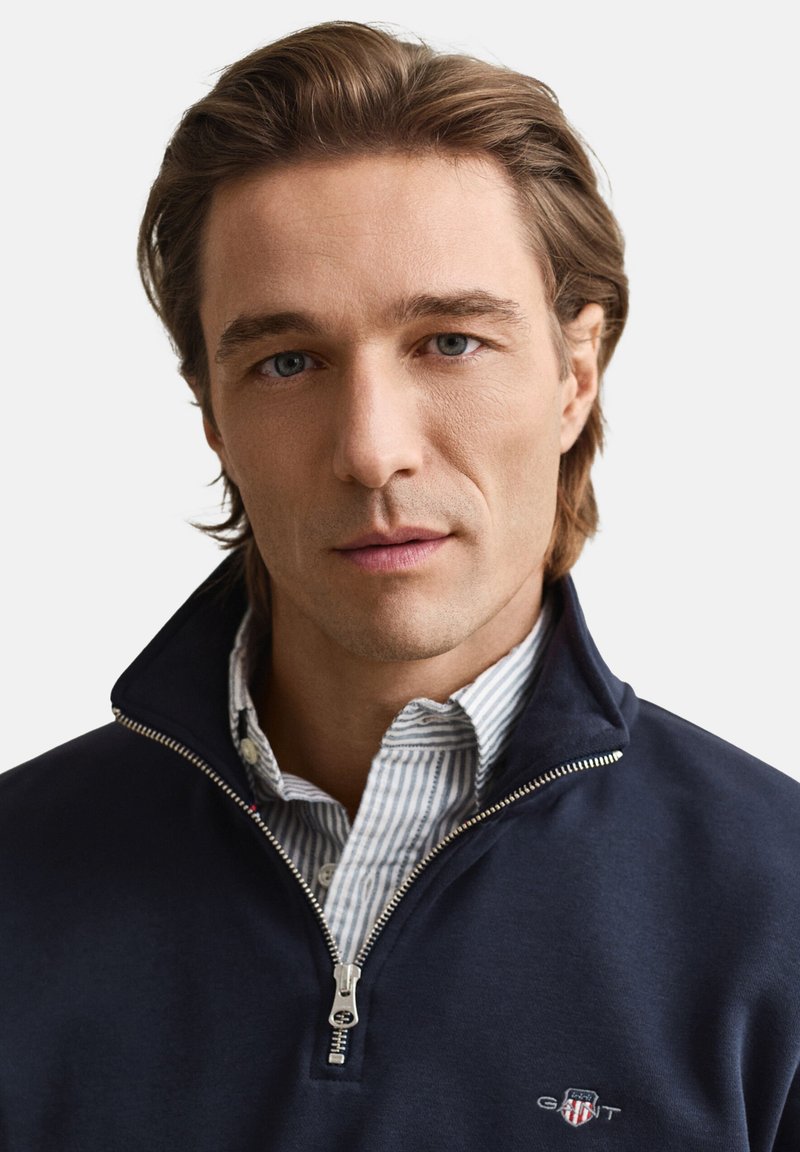 Marineblauer Pullover mit Reißverschluss und hohem Kragen, getragen über einem blau-weiß gestreiften Hemd. Mit silbernem Reißverschluss und Logo auf der linken Brust.