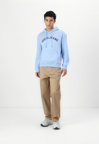 Sudadera de color azul claro con el logo de "GUESS JEANS" en negro, bolsillo canguro, puños acanalados, combinada con pantalones beige con pliegues y zapatillas gris.